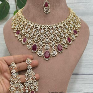 May include: A gold-toned necklace set with a matching headpiece and earrings. The jewelry features clear, sparkling stones and ruby-colored teardrop-shaped accents. The set is displayed on a mannequin.