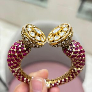 May include: A gold cuff bracelet featuring a dense pavé of deep pink stones and small clear crystals. The two ends of the open bracelet are capped with large, circular gold settings, each containing a central marquise stone surrounded by clear crystals.