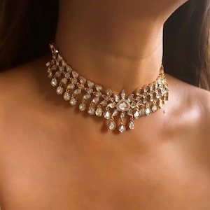 May include: A gold choker necklace adorned with clear, teardrop-shaped gemstones. The necklace has a floral centrepiece and dangling gems, creating a glamorous and elegant appearance. The jewellery is worn around the neck.