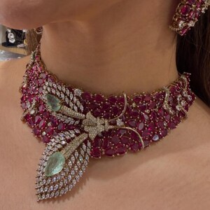 May include: A luxurious necklace and earring set featuring a vibrant array of ruby-red gemstones. The necklace showcases a central butterfly design with diamond accents and two pear-shaped green gemstones. The earrings mirror the ruby and diamond design.