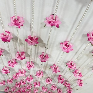 May include: A decorative ceiling display featuring strings of faux pearls and vibrant pink roses. The roses are evenly spaced along the strings, creating a visually appealing and elegant arrangement. The background is a light-colored surface.