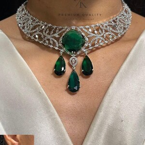 May include: A silver-toned necklace with a large emerald-green central stone and three dangling teardrop-shaped emerald-green pendants. The necklace has intricate detailing and matching earrings. The text "PREMIUM QUALITY" is visible.