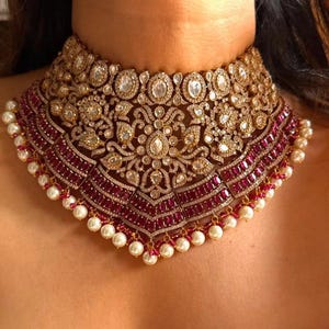 Könnte beinhalten: Ein luxuriöses, goldfarbenes Choker-Halsband mit aufwendigen Details. Die Halskette hat einen Mittelteil aus funkelnden, klaren Steinen und rubinroten Akzenten sowie eine Reihe von Perlen am unteren Rand. Ein auffälliges Schmuckstück.