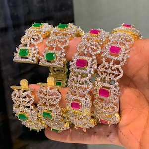May include: Gold bangles with detailed openwork patterns. The bangles are adorned with square-cut green and magenta gemstones, set amidst many small, sparkling crystals. The image shows the bangles held in a hand, highlighting their ornate design.