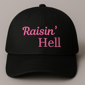Puede incluir: Gorra de camionero negra con visera curva y parte trasera de malla. La frase "Raisin' Hell" está bordada en rosa en la parte delantera. La gorra tiene un cierre a presión para ajustar la talla.