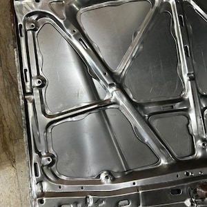 May include: A silver-colored car hood, showing the underside with structural supports and cutouts. The metal surface has a brushed appearance, with visible weld marks and mounting holes. The hood's design includes geometric shapes and reinforcing ribs.