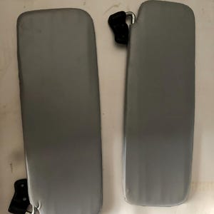 Puede incluir: Dos parasoles grises para coche. Cada parasol tiene forma rectangular con esquinas redondeadas. Los parasoles tienen herrajes de montaje negros. Los parasoles se muestran sobre un fondo blanco.