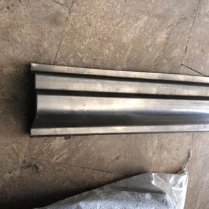May include: A long, silver metal architectural moulding with a curved, grooved design. The moulding has a smooth, reflective surface. The piece is lying on a textured, light brown surface.