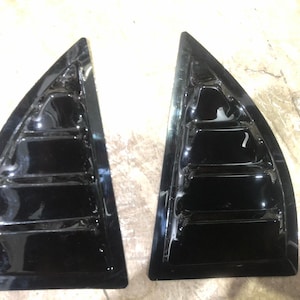 May include: A pair of glossy black car window louvres. Each louvre is triangular with horizontal slats. The louvres are designed to attach to the rear side windows of a vehicle. The finish is smooth and reflective.