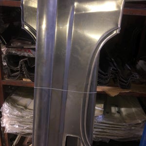 Puede incluir: Un panel de coche metálico grande, de color plateado, con forma de guardabarros curvado y un recorte rectangular. El panel tiene crestas verticales y está asegurado con una cuerda blanca. El fondo muestra estantes con más paneles metálicos.