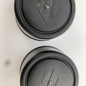 Puede incluir: Dos objetos negros y redondos con un borde circular elevado y un cierre central plateado. Cada objeto tiene un diseño en forma de diamante con una letra estilizada en el centro. Los objetos están sobre un fondo blanco.