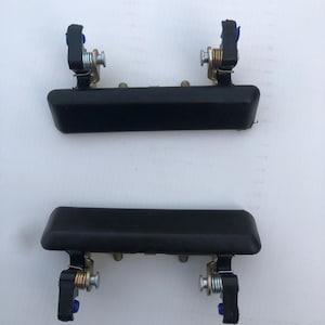 May include: Two black exterior door handles with mounting hardware. Each handle has a rectangular shape with rounded edges. The hardware includes springs, metal brackets, and blue plastic components. The handles are likely for a vehicle.