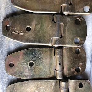 Puede incluir: Primer plano de cuatro bisagras metálicas vintage. Cada bisagra es de color plateado deslustrado con un extremo redondeado y tres agujeros. El texto "UAE 100%" está estampado en la bisagra superior. Las bisagras están conectadas en pares.