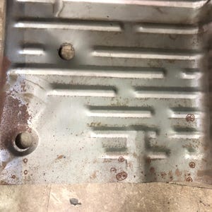 Può includere: Una lamiera metallica rettangolare e usurata con una serie di linee in rilievo parallele e perpendicolari, che creano un motivo a griglia. Il metallo è grigio argento con macchie di ruggine e due fori circolari. La lamiera sembra essere un componente di un veicolo.