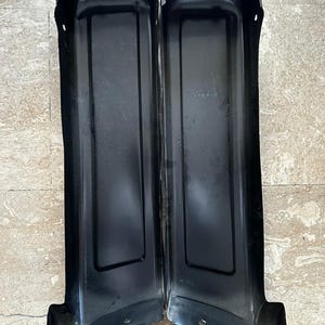May include: Two black metal panels, likely automotive parts, are positioned side-by-side. Each panel has a rectangular shape with a curved bottom and a slightly angled top. The panels appear to be made of a sturdy metal material.