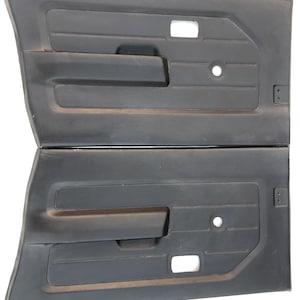 May include: Two black car door panels. Each panel features a rectangular armrest, a small circular hole, and a rectangular opening. The panels are made of a textured material, designed for a vehicle's interior.