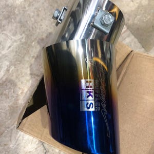 Puede incluir: Dos puntas de escape, una de acero inoxidable y otra con un acabado degradado azul y dorado. La punta inferior presenta el logotipo de HKS. Las puntas están aseguradas con abrazaderas y pernos metálicos. El diseño sugiere una mejora de rendimiento.