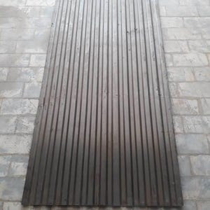 May include: A large, rectangular sheet of corrugated metal. The metal has a dark gray color and vertical ridges running its length. The sheet is lying on a brick surface.