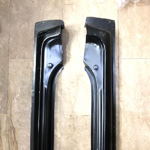 May include: Two black metal car parts, possibly trim or structural components, are displayed against a neutral background. Each piece has a curved, elongated shape with a series of parallel ridges and a rounded end. The parts appear to be new.