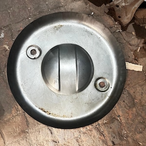 May include: A circular, silver-colored metal object with a central, raised, rounded section and two circular holes. The object appears to be a cover or lid, possibly for a mechanical component. The surface shows signs of wear and use.