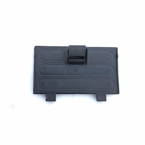 Puede incluir: Una tapa de compartimento de batería rectangular de plástico negro con un pestillo con bisagras. La tapa tiene tres secciones elevadas de forma ovalada y dos pequeñas pestañas en la parte inferior. La tapa está diseñada para proteger las baterías.