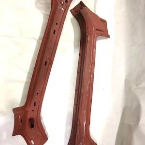 Puede incluir: Dos componentes de bastidor de coche de metal de color marrón rojizo. Uno es una pieza larga y recta con varios agujeros y una base acampanada. El otro es curvo con un color y una construcción similares. Ambas parecen piezas usadas o recuperadas.