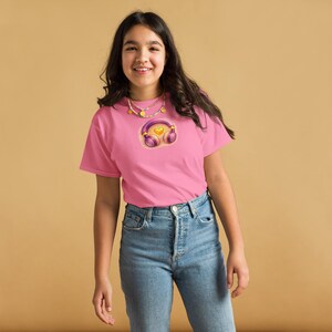 May include: Pink t-shirt featuring a graphic of purple headphones with a yellow heart in the center. The headphones are set against a light pink circle. The person is wearing blue jeans and a gold necklace.