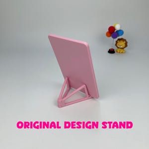 May include: A pink rectangular stand with a triangular support. The stand is angled back, and the text "ORIGINAL DESIGN STAND" is displayed in bright pink at the bottom of the image. A small lion figurine and a cluster of balloons are in the background.