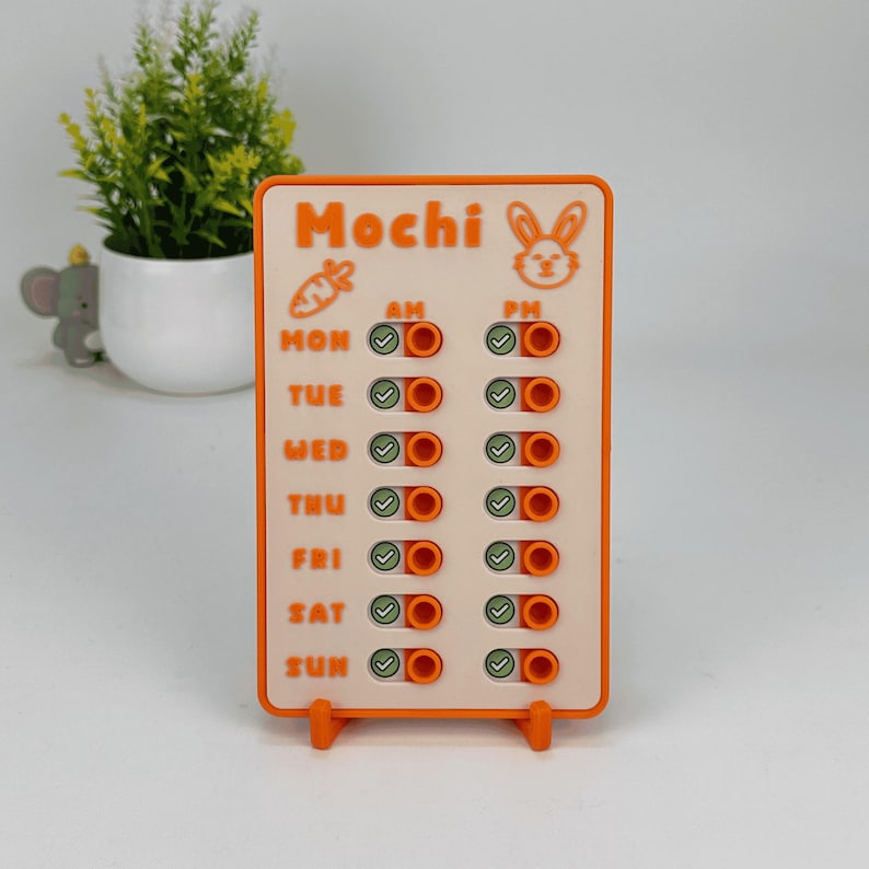 May include: An orange and white weekly planner with the word "Mochi" at the top, along with a carrot and bunny graphic. The planner has toggle switches for AM and PM, with days of the week listed. The planner is standing on a small orange stand.