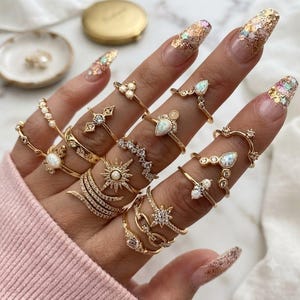 May include: A collection of gold-toned rings worn on fingers, featuring various designs including starbursts, teardrop-shaped opals, pavé-set crystals, delicate chains, and thin bands with small spherical accents, all showcasing a metallic finish.
