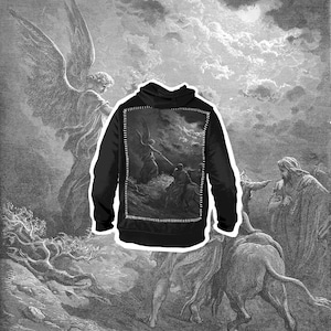 Peut inclure: Sweat à capuche noir avec une illustration monochrome détaillée au dos, représentant une scène dramatique avec des personnages et des nuages. Le sweat à capuche est placé sur un fond d'œuvre d'art similaire.