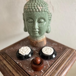 Könnte beinhalten: Eine grüne Buddha-Kopf-Statue mit geschlossenen Augen. Zwei weiße Blumen- und Kreisdesigns auf schwarzen Sockeln und ein kleines Holzobjekt werden auf einer Marmoroberfläche ausgestellt.