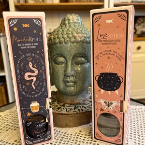 Könnte beinhalten: Zwei Raumduftdiffusoren in dekorativen Schachteln. Einer trägt die Aufschrift "Moondust Spell" mit einer Schlangenillustration, der andere "Love Potion" mit einem Kessel und einem Schmetterling. Beide haben eine Glasflasche mit Stäbchen.