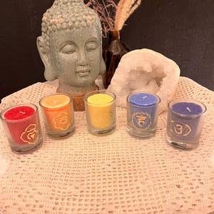 Könnte beinhalten: Fünf kleine Glas-Votivkerzen mit farbigem Wachs: rot, orange, gelb, blau und lila. Jede Kerze hat ein Chakra-Symbol. Ein hellgrüner Buddha-Kopf und eine weiße Geode befinden sich im Hintergrund.