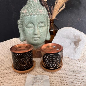 Könnte beinhalten: Eine ruhige Komposition mit einem hellgrünen Buddha-Kopf aus Keramik, zwei kupfer-schwarzen Kerzenhaltern mit aufwendigen Designs und einer weißen Geode. Die Kerzenhalter stehen auf Holzsockeln. Ein kleines Päckchen mit Text ist ebenfalls vorhanden.