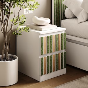Puede incluir: Mesita de noche blanca con dos cajones, acentuada con tiras verticales de musgo verde y madera clara. Un cuenco de cerámica blanco y un libro descansan encima. Una planta en maceta está en primer plano.