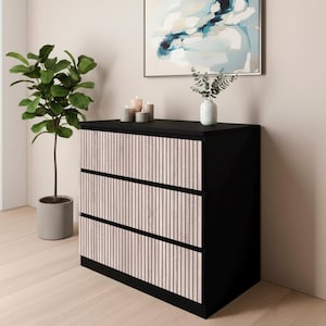 May include: A black three-drawer chest of drawers with light-coloured vertical wooden slats on the drawer fronts. The room includes a potted plant, candles, and an abstract painting in a frame.