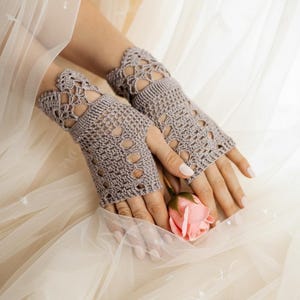 May include: A pair of gray, open-work crochet fingerless gloves. The gloves have a scalloped edge and are made of a soft, textured yarn. A pink rose is held between the hands. The background is a soft, neutral color.