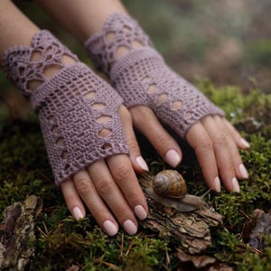 Puede incluir: Un par de guantes sin dedos de color malva con un intrincado patrón de ganchillo, que presenta un borde festoneado en la muñeca y un delicado diseño calado a lo largo de la mano, ofreciendo una apariencia texturizada.