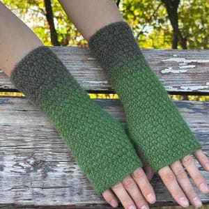 Peut inclure: Une paire de mitaines en crochet avec un motif texturé. Les gants présentent un dégradé allant d'un vert olive foncé au niveau du poignet vers un vert herbe plus clair et vif vers les doigts, couvrant l'avant-bras tout en laissant les doigts libres.