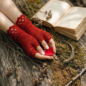 Puede incluir: Un par de guantes sin dedos rojos con un borde festoneado. Los guantes están hechos a ganchillo y se exhiben sobre una superficie de madera con un libro y un pétalo rojo. Los guantes son de un tono rojo vibrante.