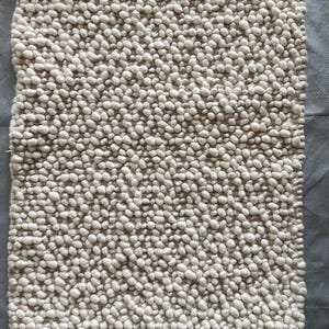 May include: A rectangular area rug featuring a thick, high-pile texture composed of numerous small, rounded, off-white wool-like knots densely packed together across the entire surface, creating a uniform, bumpy, and tactile appearance.