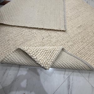 May include: A cream-colored area rug featuring a textured surface composed of numerous small, rounded felted wool balls. The rug is folded over to reveal a flat-woven underside with a matching cream-colored fabric border along the edges.
