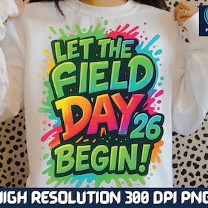 May include: White sweatshirt with a colorful graphic that reads "LET THE FIELD DAY 26 BEGIN!" The design features vibrant green, yellow, orange, and pink text with a splash effect. The sweatshirt is a crew neck style.
