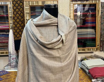 May include: A neutral-toned shawl draped over a mannequin. The shawl has a subtle sheen and a delicate, silver-colored trim along the edges. The background features shelves displaying various textiles, suggesting a textile shop or boutique.
