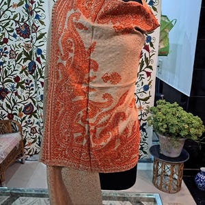 Op de afbeelding: Een beige sjaal met ingewikkelde oranje borduursels. De sjaal is over een mannequin gedrapeerd en toont de gedetailleerde paisley- en bloempatronen. Het ontwerp heeft een franje langs de onderrand.