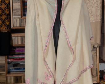 May include: A cream-colored shawl with a pink and black embroidered border and accents. The shawl is draped over a black mannequin. The background includes a wooden cabinet with shelves displaying various textiles.
