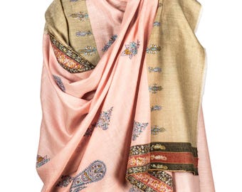 May include: A pink and tan shawl with intricate floral and paisley patterns. The shawl is draped over a mannequin, showcasing its design and texture. The edges feature a detailed border with various colors.