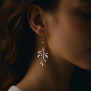 Può includere: Orecchino argentato con un design a ramo e piccoli dettagli di foglie. L'orecchino pende da un semplice gancio, mostrando un'estetica minimalista e ispirata alla natura. L'orecchino è mostrato su uno sfondo sfocato.