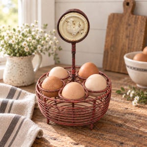 May include: A round, red, wire egg basket with a central handle topped by a circular metal plaque featuring a raised white rooster design. The basket holds five brown and cream-coloured eggs in individual wire rings, supported by small feet.